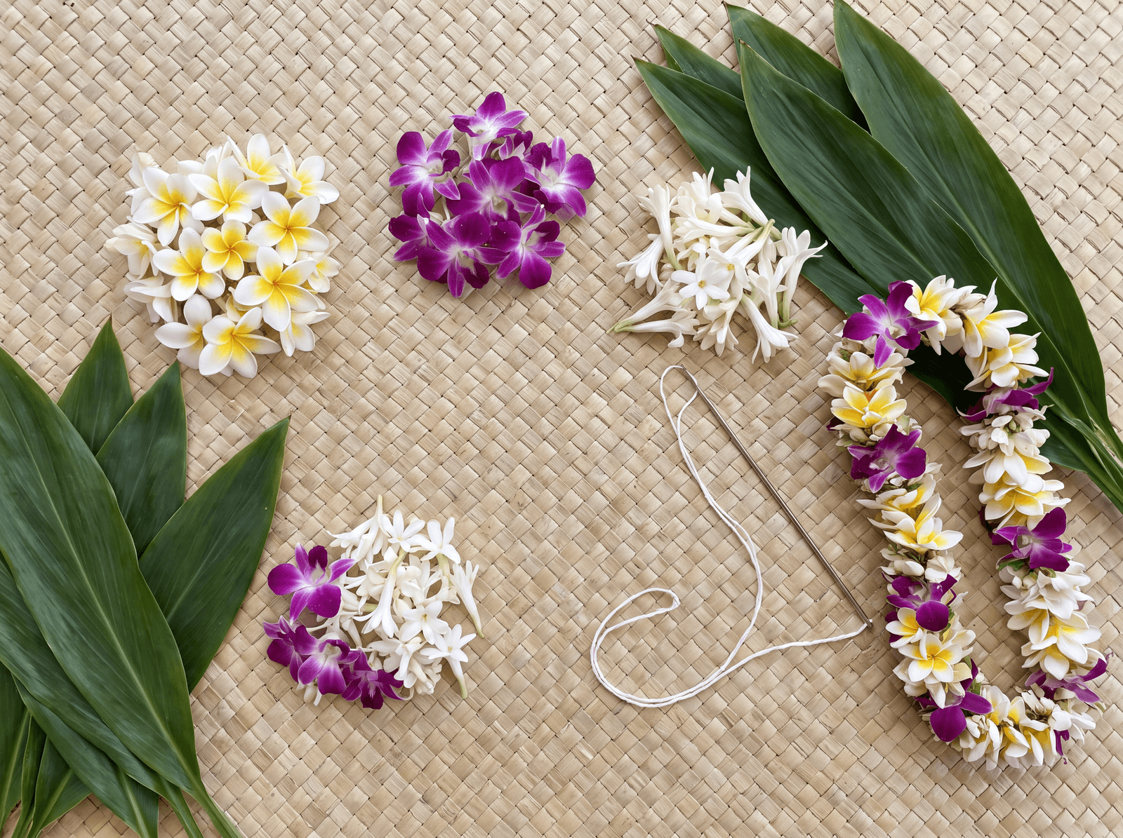 Lei-making flowers, ti leaves and a lei needle on a lauhala mat